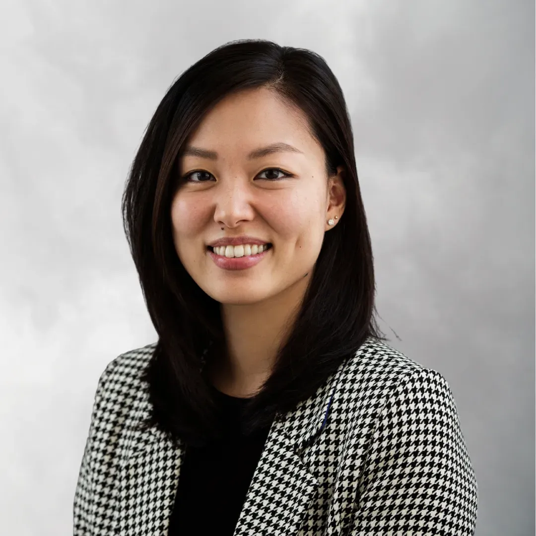 A woman with shoulder-length dark hair smiling, wearing a black and white houndstooth blazer and small stud earrings against a cloudy gray background.