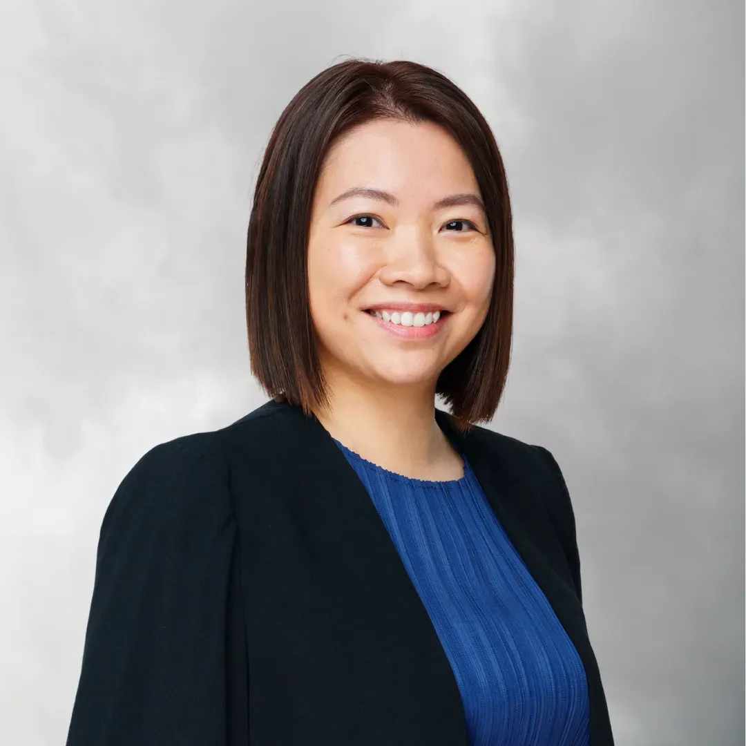 Professional woman with shoulder-length brown hair, smiling, wearing a black blazer over a blue top, against a gray background.