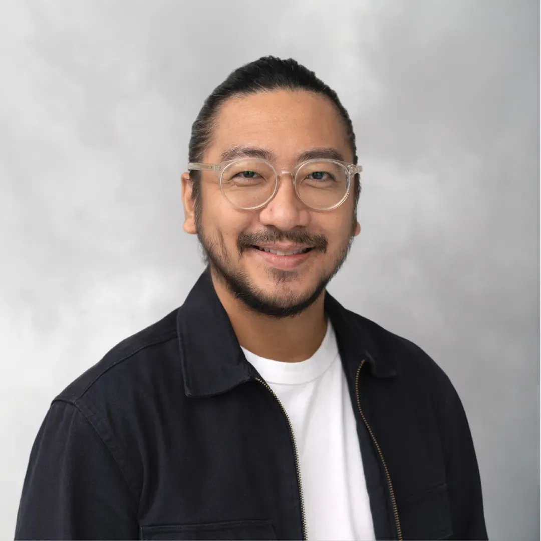 A man with glasses, a beard, and mustache smiling at the camera, wearing a black jacket over a white t-shirt with a gray cloudy sky background.