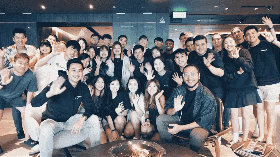 Group of young adults posing for a photo indoors, smiling, some waving, with a mix of men and women in casual and semi-formal attire.
