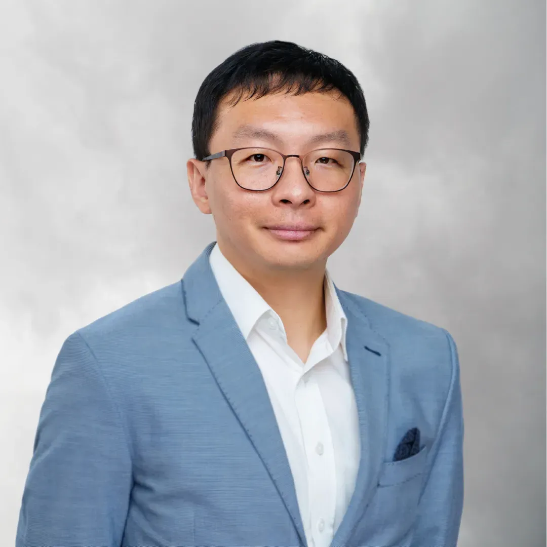 Professional portrait of a man wearing glasses, a light blue blazer, and a white dress shirt, smiling slightly against a gray background.